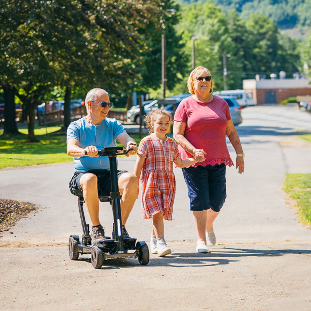 Lifestyle shot of a man riding the black Pride Go Go Carbon scooter on a paved path while enjoying an active outing with his family.