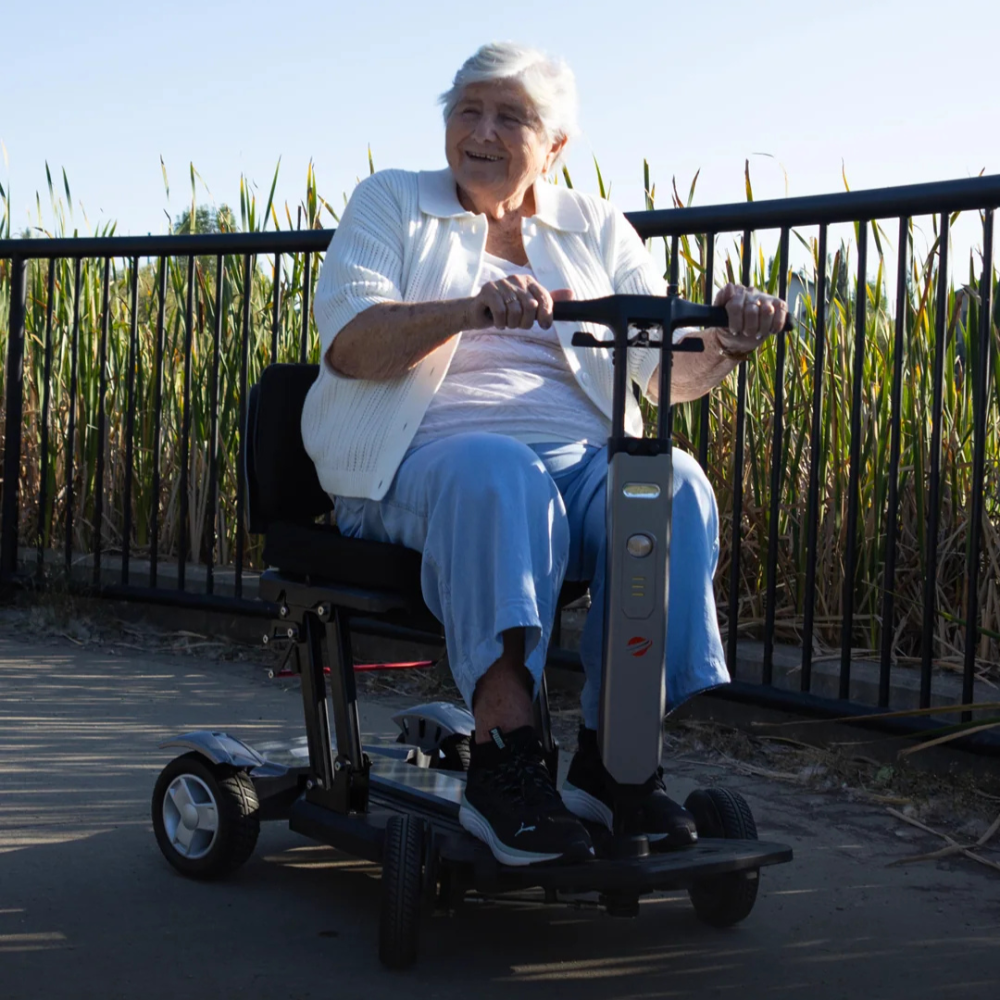 A woman riding the Travel Buggy HELIX mobility scooter along a paved outdoor path, Care Made