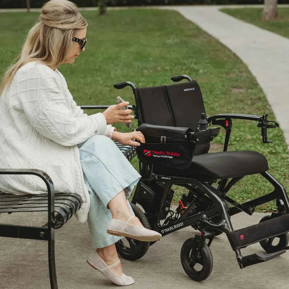 Person sitting comfortably in a power wheelchair outdoors with the Travel Buggy armrest organizer attached for easy access.