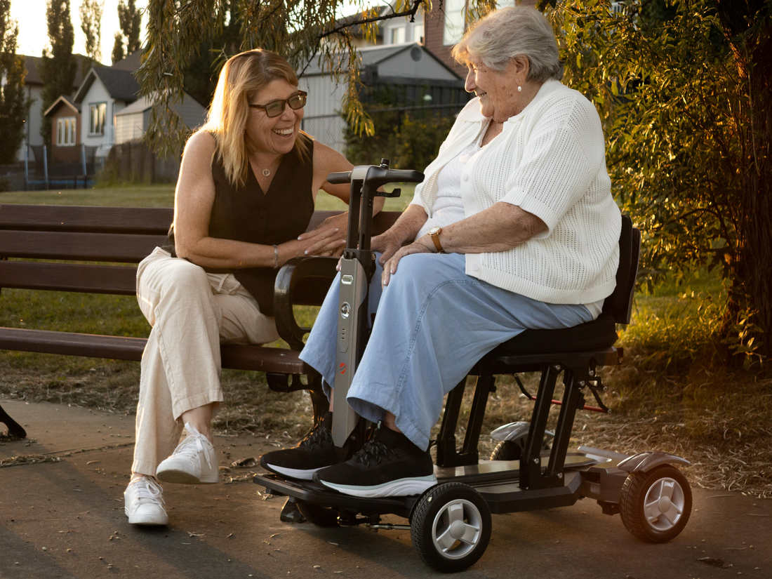 Older woman on a Travel Buggy HELIX scooter chatting with a younger woman on a park bench