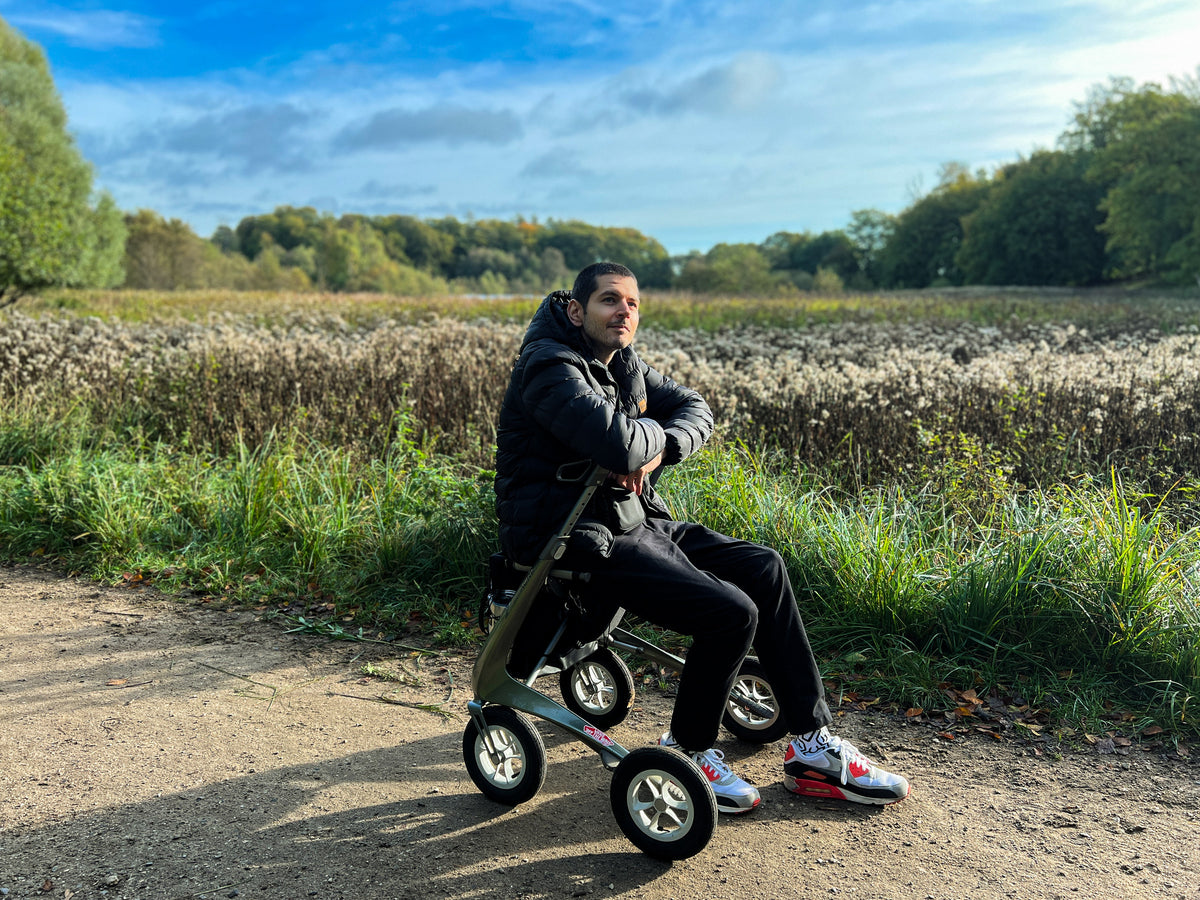 Man sitting on the seat of a Defender Green byACRE Carbon Overland rollator on a dirt path beside a field