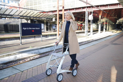 Woman walking through a train station with an Oyster White byACRE Carbon Ultralight rollator
