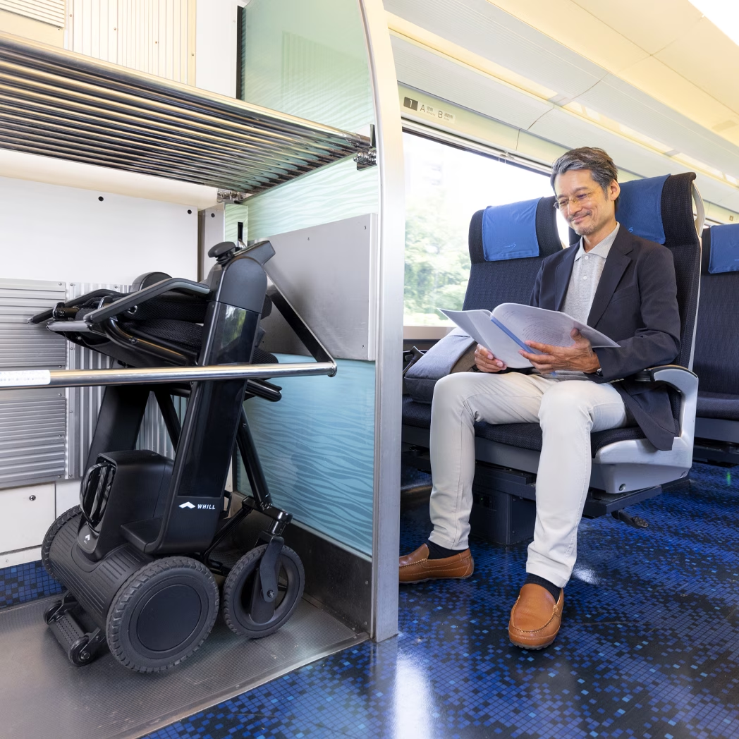 Folded WHILL Model F power wheelchair stored beside a seated passenger on a train