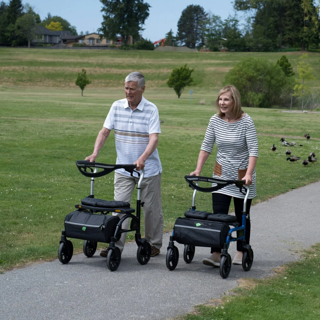A couple enjoying a sunny outdoor walk together, with the woman comfortably utilizing the pink Evolution Trillium rollator.