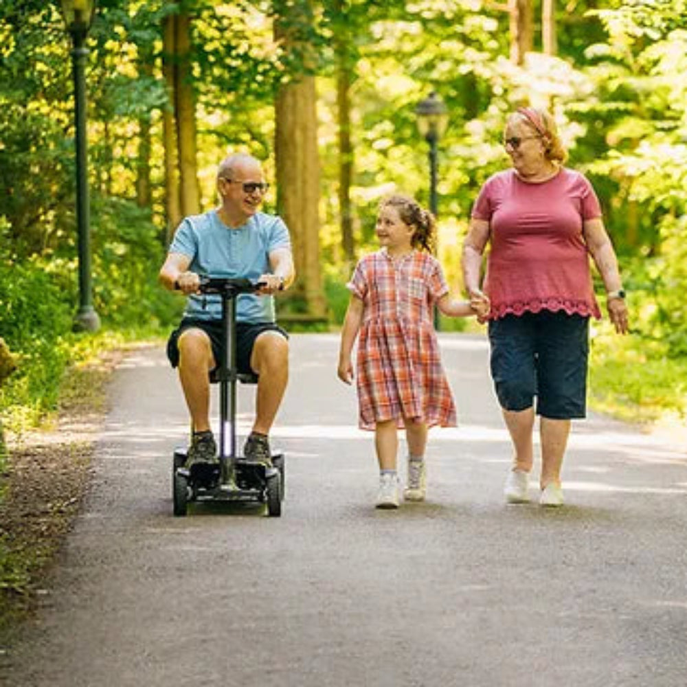 Lifestyle shot of a man comfortably driving the yellow Pride Go Go Carbon scooter alongside a woman walking on a park path.