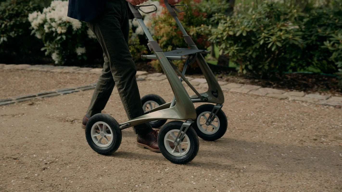 A user walking comfortably with the byACRE Carbon Overland rollator over uneven urban cobblestone streets.
