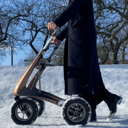 A person wearing a winter coat walking securely through snowy, icy outdoor conditions using the all-weather byACRE Carbon Overland rollator.