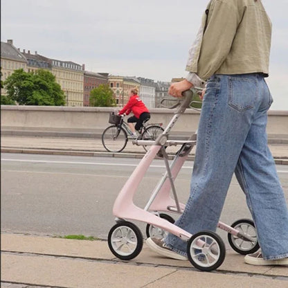 A user walking along a paved park path utilizing the lightweight pink byACRE Carbon Ultralight rollator for support.