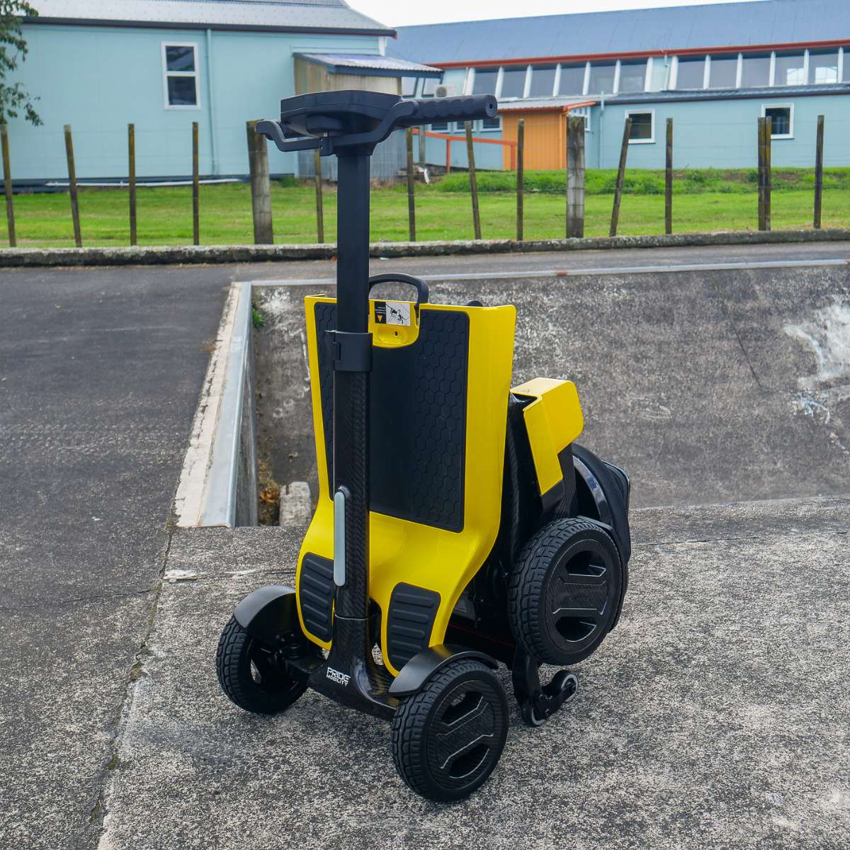 Front angled view of the yellow Pride Go Go Carbon mobility scooter parked outdoors, showcasing its sleek travel-ready design.