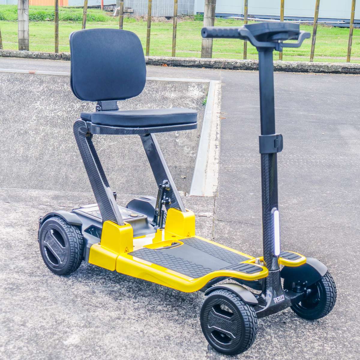 The bright yellow Pride Mobility Go Go Carbon lightweight travel scooter sitting on an outdoor paved path ready for everyday mobility.