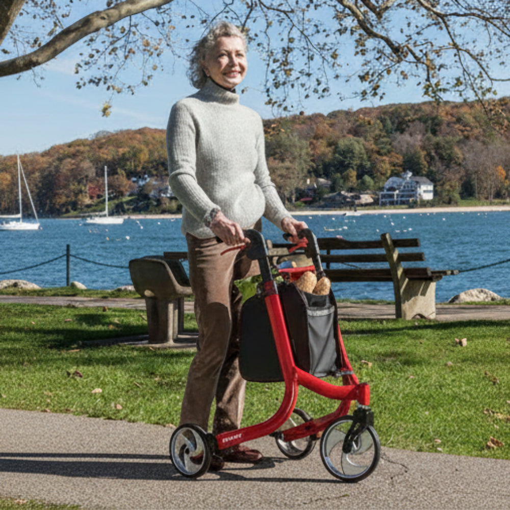 Older person standing with a Red Drive Medical Nitro three-wheel rollator beside a marina