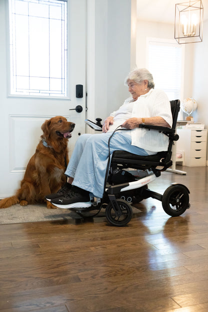 Woman seated in a white Travel Buggy Grand Tourer GT power wheelchair beside a dog at home