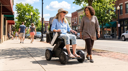 Woman using a WHILL Model C2 High Back power wheelchair with white arm covers on a sidewalk beside a companion