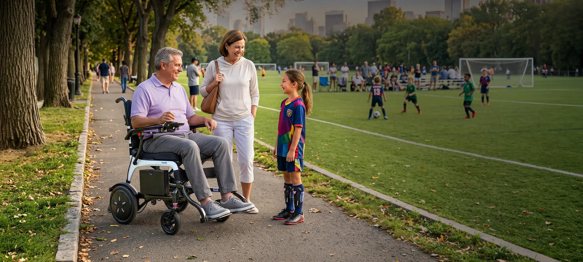 Man seated in a WHILL MODO Breeze power wheelchair with a white frame beside a woman and child near a soccer field