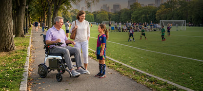 Man seated in a WHILL MODO Breeze power wheelchair with a white frame beside a woman and child near a soccer field