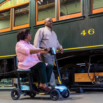 Lifestyle image of an older couple in the sidewalk, with the woman comfortably driving the blue Pride Go-Go Super Portable scooter.